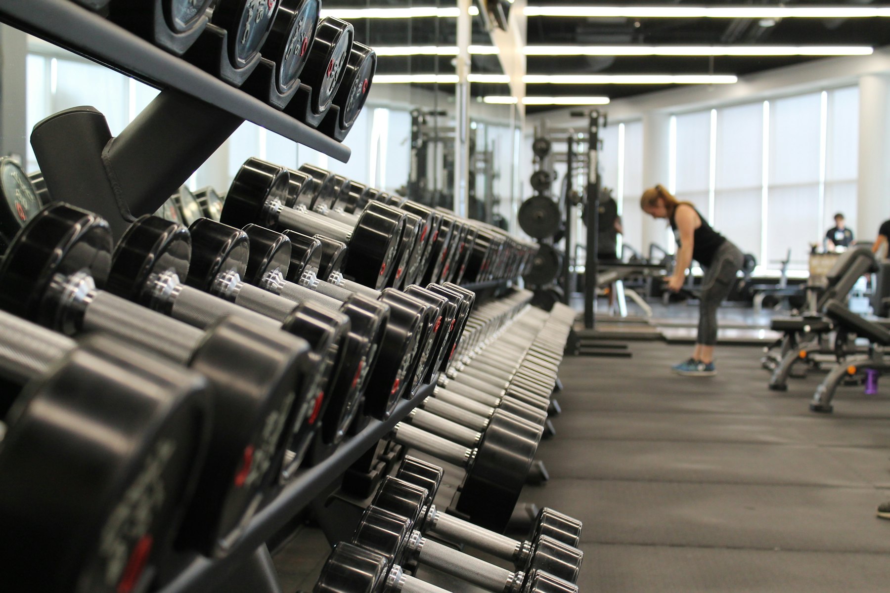 Rows of dumbbells on a rack, with a person training further back on a gym floor